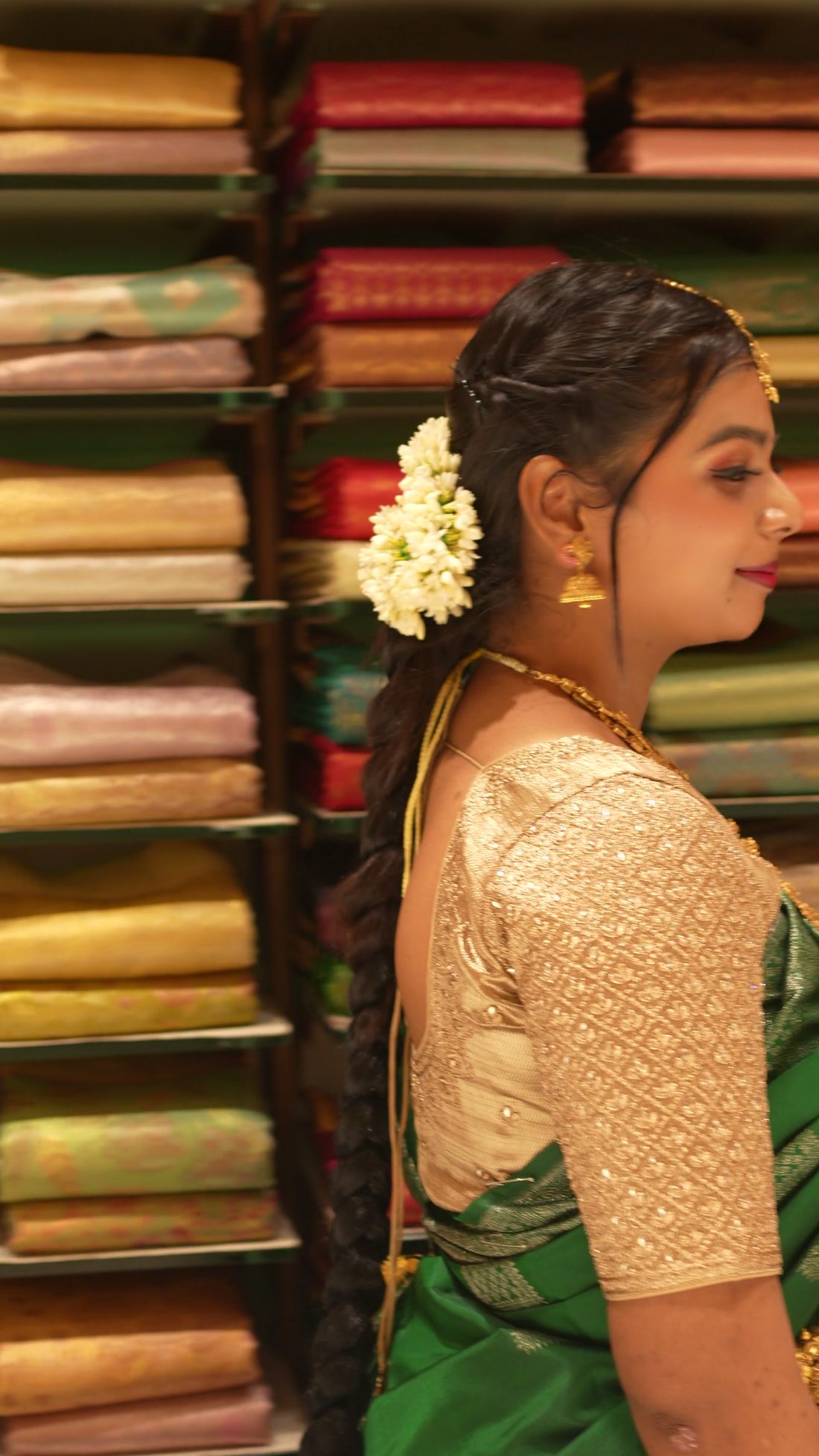 Woman in a green Kanchipuram silk saree with gold zari border, standing before shelves of folded silk sarees in a saree shop