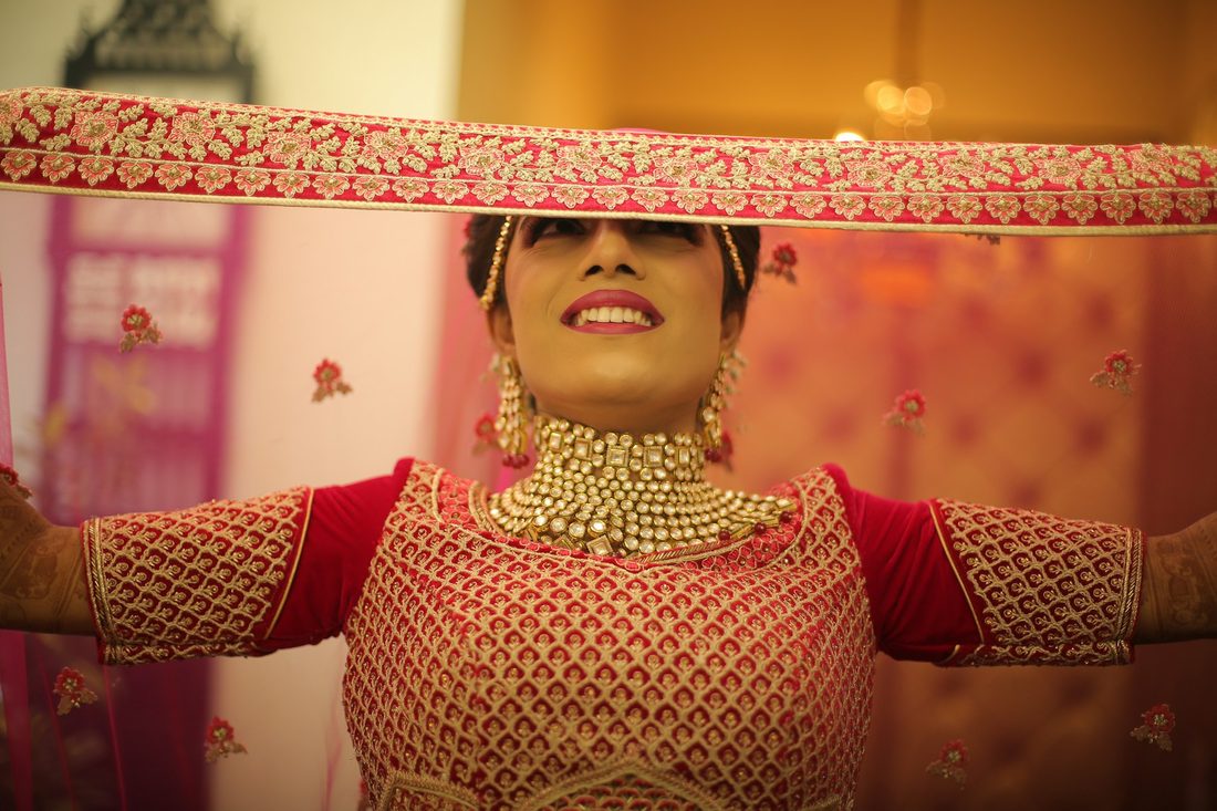 Bride in a red and gold embroidered saree lifting her veil, smiling