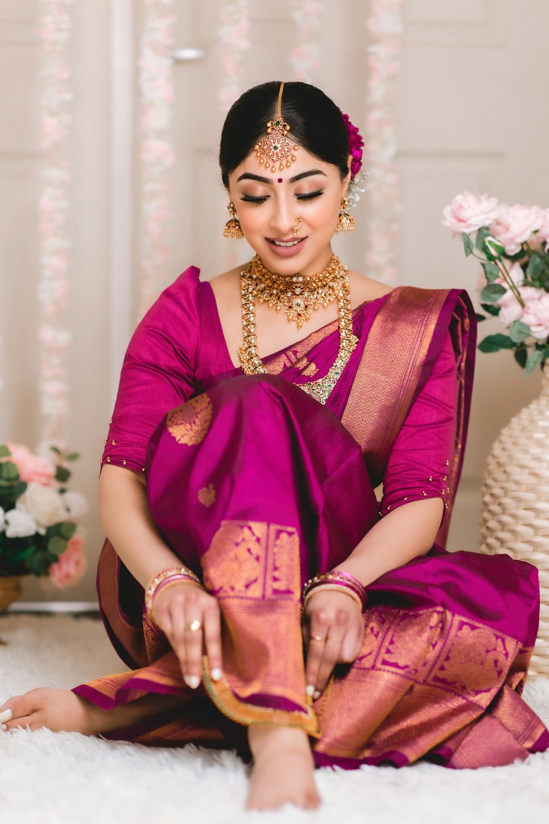 Woman seated in a magenta silk saree with gold zari, surrounded by flowers